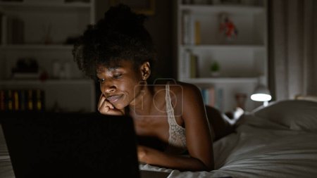 Téléchargez les photos : Femme afro-américaine portant de la lingerie à l'aide d'un ordinateur portable couché sur le lit dans la chambre - en image libre de droit