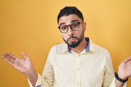 Photo for Hispanic young man wearing business clothes and glasses clueless and confused expression with arms and hands raised. doubt concept. - Royalty Free Image