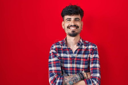 Téléchargez les photos : Jeune homme hispanique avec la barbe debout sur fond rouge heureux visage souriant avec les bras croisés regardant la caméra. personne positive. - en image libre de droit