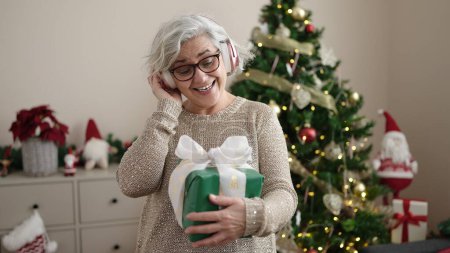 Photo for Middle age woman with grey hair listening to music standing by christmas tree at home - Royalty Free Image