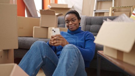 Photo for African american woman using smartphone sitting on floor at new home - Royalty Free Image