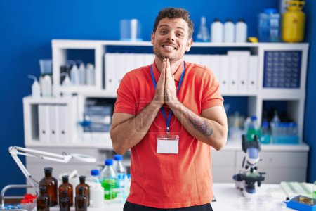 Photo for Young hispanic man working at scientist laboratory praying with hands together asking for forgiveness smiling confident. - Royalty Free Image