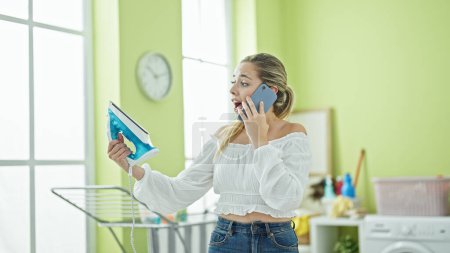 Téléchargez les photos : Jeune femme blonde parlant sur smartphone regardant machine à repasser surpris à la buanderie - en image libre de droit