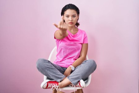 Photo for Hispanic young woman sitting on chair over pink background showing middle finger, impolite and rude fuck off expression - Royalty Free Image