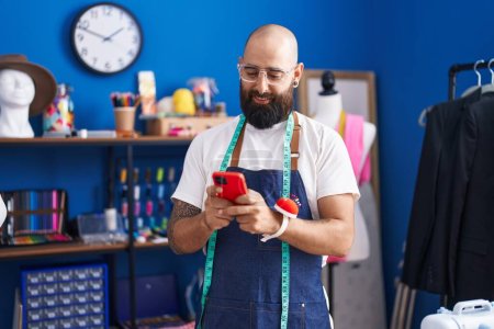 Téléchargez les photos : Jeune homme chauve tailleur souriant confiant en utilisant smartphone à l'usine de vêtements - en image libre de droit