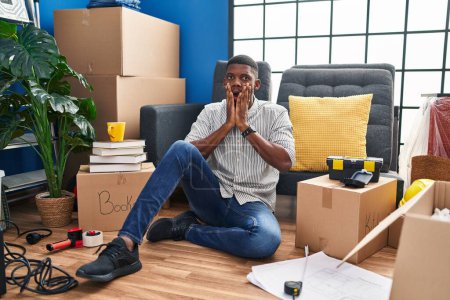 Foto de Hombre afroamericano sentado en el suelo en el nuevo hogar asustado y sorprendido, sorpresa y expresión asombrada con las manos en la cara - Imagen libre de derechos