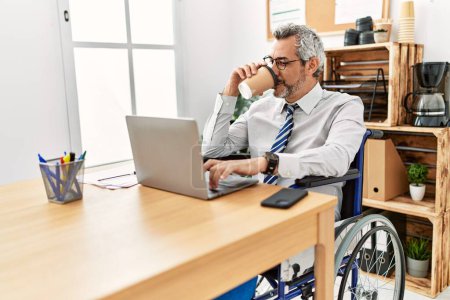 Foto de Hombre de pelo gris de mediana edad trabajador de negocios sentado en silla de ruedas bebiendo café en la oficina - Imagen libre de derechos