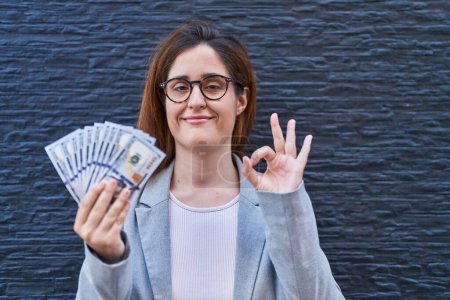 Téléchargez les photos : Brunette femme tenant des dollars faisant signe ok avec les doigts, sourire geste amical excellent symbole - en image libre de droit