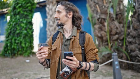 Téléchargez les photos : Jeune homme hispanique touriste en utilisant smartphone tenant café au parc - en image libre de droit