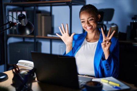 Foto de Hermosa mujer afroamericana que trabaja en la oficina por la noche mostrando y señalando hacia arriba con los dedos número ocho mientras sonríe confiada y feliz. - Imagen libre de derechos
