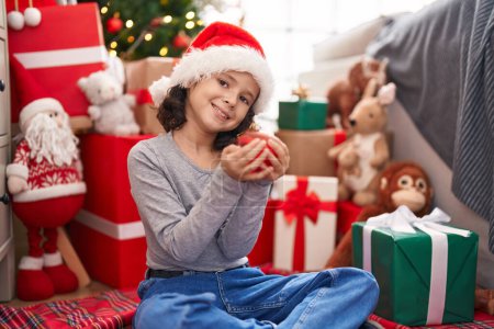Photo for Adorable chinese girl holding ball decoration sitting on floor by christmas tree at home - Royalty Free Image