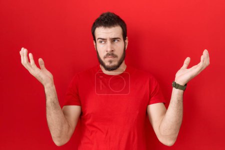 Photo for Young hispanic man wearing casual red t shirt clueless and confused expression with arms and hands raised. doubt concept. - Royalty Free Image