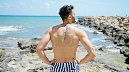 Téléchargez les photos : Jeune homme hispanique touriste portant maillot de bain debout vers l'arrière à la plage - en image libre de droit