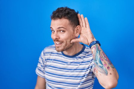 Photo for Young hispanic man standing over blue background smiling with hand over ear listening an hearing to rumor or gossip. deafness concept. - Royalty Free Image