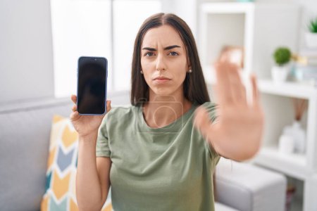 Foto de Joven morena sosteniendo smartphone mostrando pantalla en blanco con la mano abierta haciendo stop sign con expresión seria y segura, gesto de defensa - Imagen libre de derechos