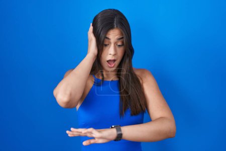 Téléchargez les photos : Femme hispanique debout sur fond bleu regardant le temps de la montre inquiet, peur de se faire tard - en image libre de droit