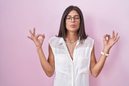 Téléchargez les photos : Brunette jeune femme debout sur fond rose portant des lunettes détendues et souriantes avec les yeux fermés faisant geste de méditation avec les doigts. concept de yoga. - en image libre de droit