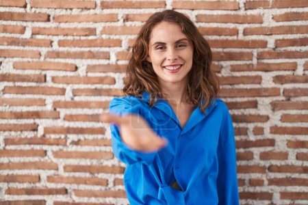 Téléchargez les photos : Belle femme brune debout sur un mur de briques souriant joyeuse offrant main de paume donnant assistance et acceptation. - en image libre de droit