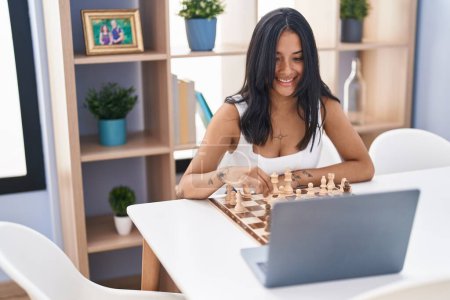 Téléchargez les photos : Jeune femme hispanique jouant jeu d'échecs en ligne assis sur la table à la maison - en image libre de droit