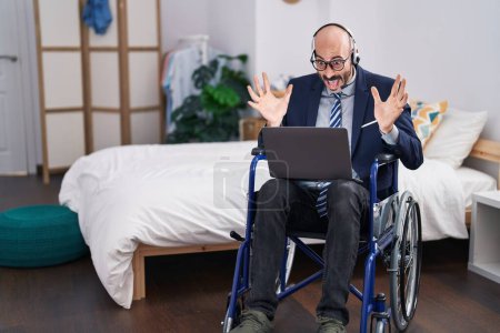 Téléchargez les photos : Homme hispanique avec barbe assis sur fauteuil roulant faire appel vidéo d'affaires célébrant la victoire avec sourire heureux et expression gagnante avec les mains levées - en image libre de droit