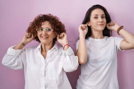 Photo for Hispanic mother and daughter wearing casual white t shirt over pink background smiling pulling ears with fingers, funny gesture. audition problem - Royalty Free Image