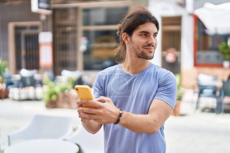 Téléchargez les photos : Jeune homme hispanique souriant confiant en utilisant un smartphone dans la rue - en image libre de droit