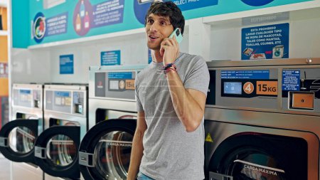 Téléchargez les photos : Jeune homme hispanique parlant sur smartphone s'appuyant sur la machine à laver souriant à la laverie - en image libre de droit
