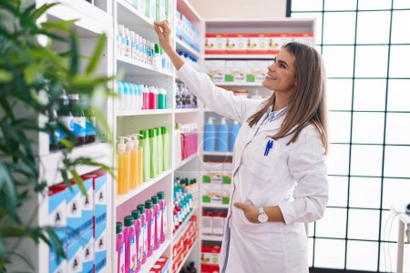 Foto de Joven hermosa mujer hispana farmacéutica sonriendo confiado sosteniendo producto en estanterías en la farmacia - Imagen libre de derechos