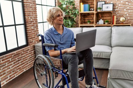 Téléchargez les photos : Jeune homme blond utilisant un ordinateur portable assis sur un fauteuil roulant à la maison - en image libre de droit