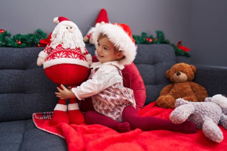 Photo for Adorable blonde toddler playing with santa claus doll sitting on sofa by christmas decoration at home - Royalty Free Image