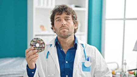 Photo for Young hispanic man nutritionist holding doughnut with serious face at clinic - Royalty Free Image