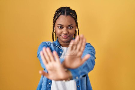 Téléchargez les photos : Femme afro-américaine avec des tresses debout sur fond jaune expression de rejet croisant les bras et les paumes faisant signe négatif, visage en colère - en image libre de droit