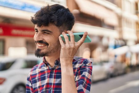 Téléchargez les photos : Jeune homme hispanique souriant confiant écoute message audio par le smartphone à la rue - en image libre de droit
