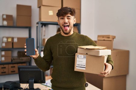 Téléchargez les photos : Arabe homme avec barbe travaillant à la petite entreprise e-commerce tenant des paquets de livraison coller la langue heureux avec une expression drôle. - en image libre de droit