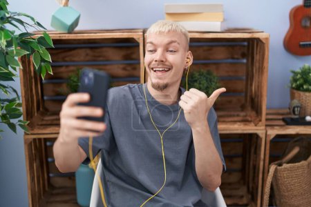 Photo for Young caucasian man doing video call with smartphone pointing thumb up to the side smiling happy with open mouth - Royalty Free Image