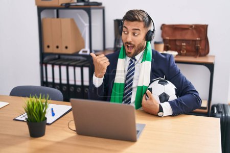 Téléchargez les photos : Bel homme hispanique regarder match de football au bureau pointant pouce vers le côté souriant heureux avec la bouche ouverte - en image libre de droit