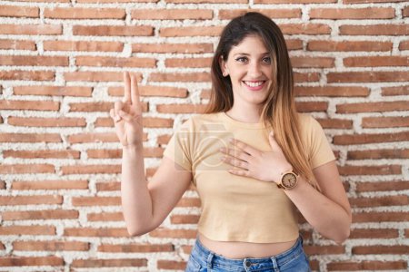 Photo for Young brunette woman standing over bricks wall smiling swearing with hand on chest and fingers up, making a loyalty promise oath - Royalty Free Image