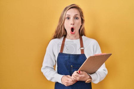 Photo for Young blonde woman wearing professional waitress apron holding clipboard scared and amazed with open mouth for surprise, disbelief face - Royalty Free Image