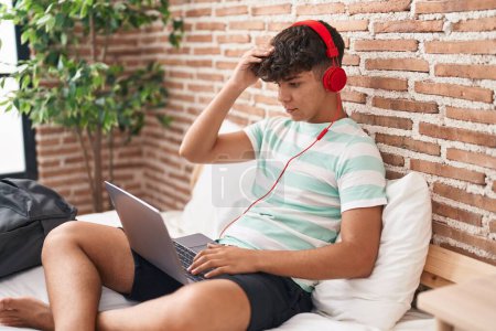 Téléchargez les photos : Adolescent hispanique utilisant un ordinateur portable à la maison assis sur la chambre à coucher stressé et frustré avec la main sur la tête, visage surpris et en colère - en image libre de droit