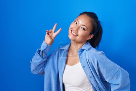 Téléchargez les photos : Asiatique jeune femme debout sur fond bleu souriant en regardant la caméra montrant les doigts faisant signe de victoire. numéro deux. - en image libre de droit