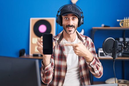 Téléchargez les photos : Homme hispanique avec barbe au studio de musique montrant smartphone souriant heureux pointant avec la main et le doigt - en image libre de droit