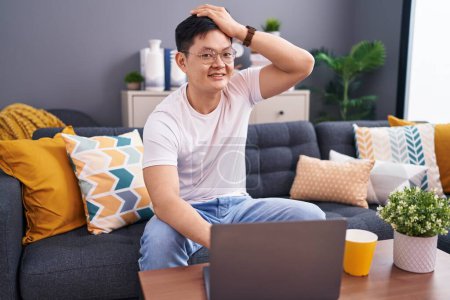 Foto de Joven hombre asiático usando el ordenador portátil en casa sentado en el sofá sonriendo seguro de tocar el pelo con la mano hacia arriba gesto, posando atractivo y de moda - Imagen libre de derechos