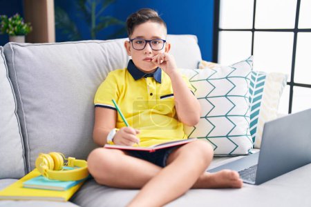 Téléchargez les photos : Jeune enfant hispanique faisant ses devoirs assis sur le canapé bouche et lèvres fermées comme zip avec les doigts. secret et silencieux, tabou parlant - en image libre de droit