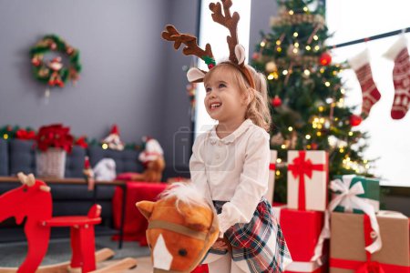 Foto de Adorable chica rubia jugando con juguete de caballo de pie junto al árbol de Navidad en casa - Imagen libre de derechos