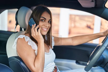 Téléchargez les photos : Young beautiful hispanic woman talking on smartphone sitting on car at street - en image libre de droit
