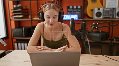 Téléchargez les photos : Jeune journaliste de radio blonde parlant avec des écouteurs et un ordinateur portable au studio de podcast - en image libre de droit