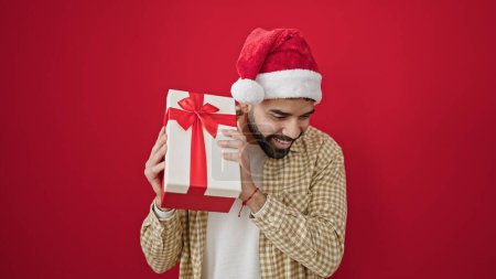 Téléchargez les photos : Jeune homme hispanique souriant confiant portant chapeau de Noël ouïe cadeau son sur fond rouge isolé - en image libre de droit