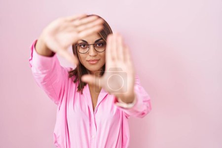 Téléchargez les photos : Jeune femme hispanique portant des lunettes debout sur fond rose faisant cadre en utilisant les paumes des mains et des doigts, perspective de la caméra - en image libre de droit
