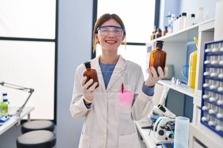 Téléchargez les photos : Jeune femme blonde scientifique tenant des bouteilles avec un sourire liquide au laboratoire - en image libre de droit