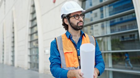 Photo for Young hispanic man architect holding blueprints looking around at construction place - Royalty Free Image